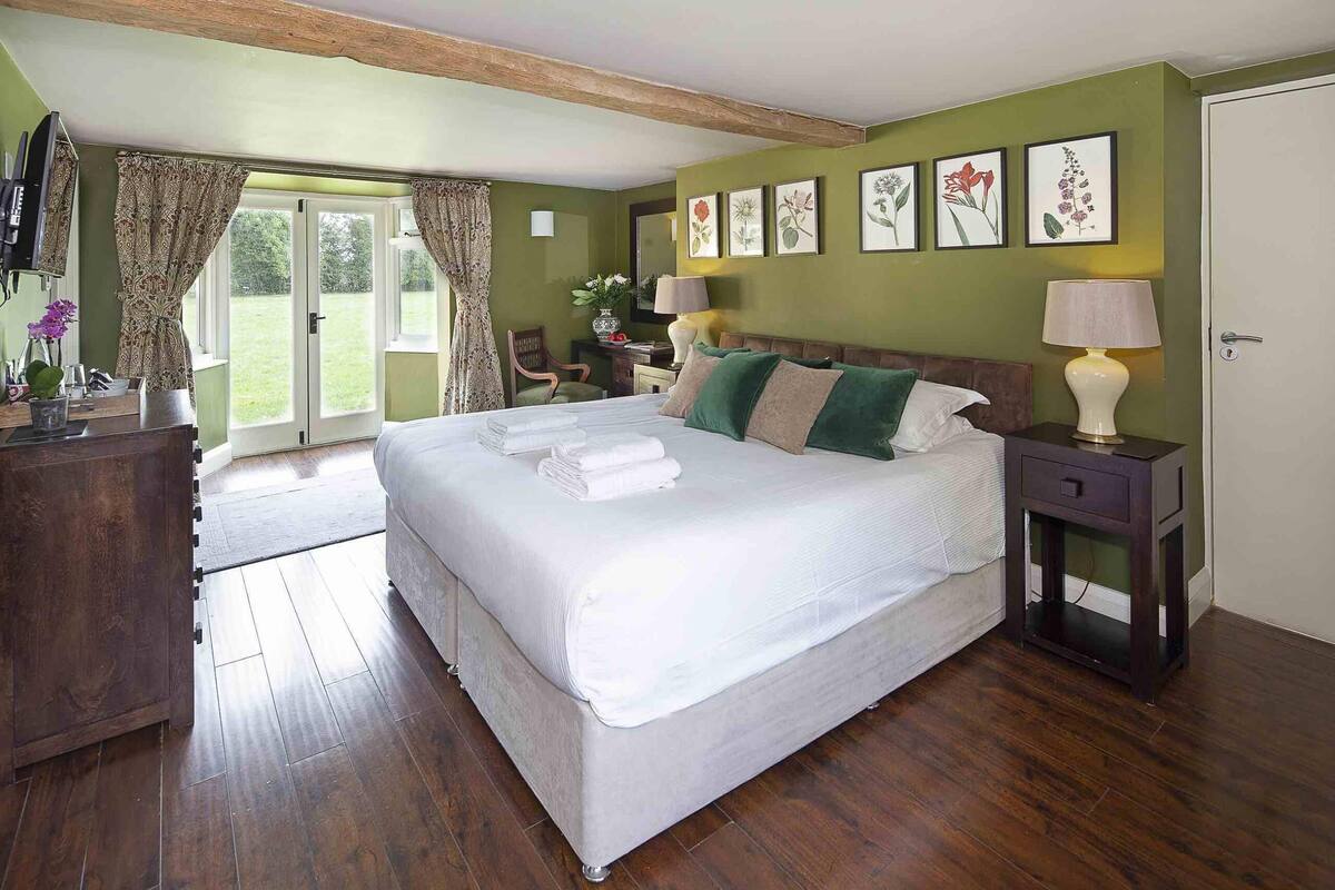 Un dormitorio con una cama grande, un aparador de madera, una mesita de noche y una silla. Hay obras de arte con motivos florales en las paredes y una puerta que da a un balcón.