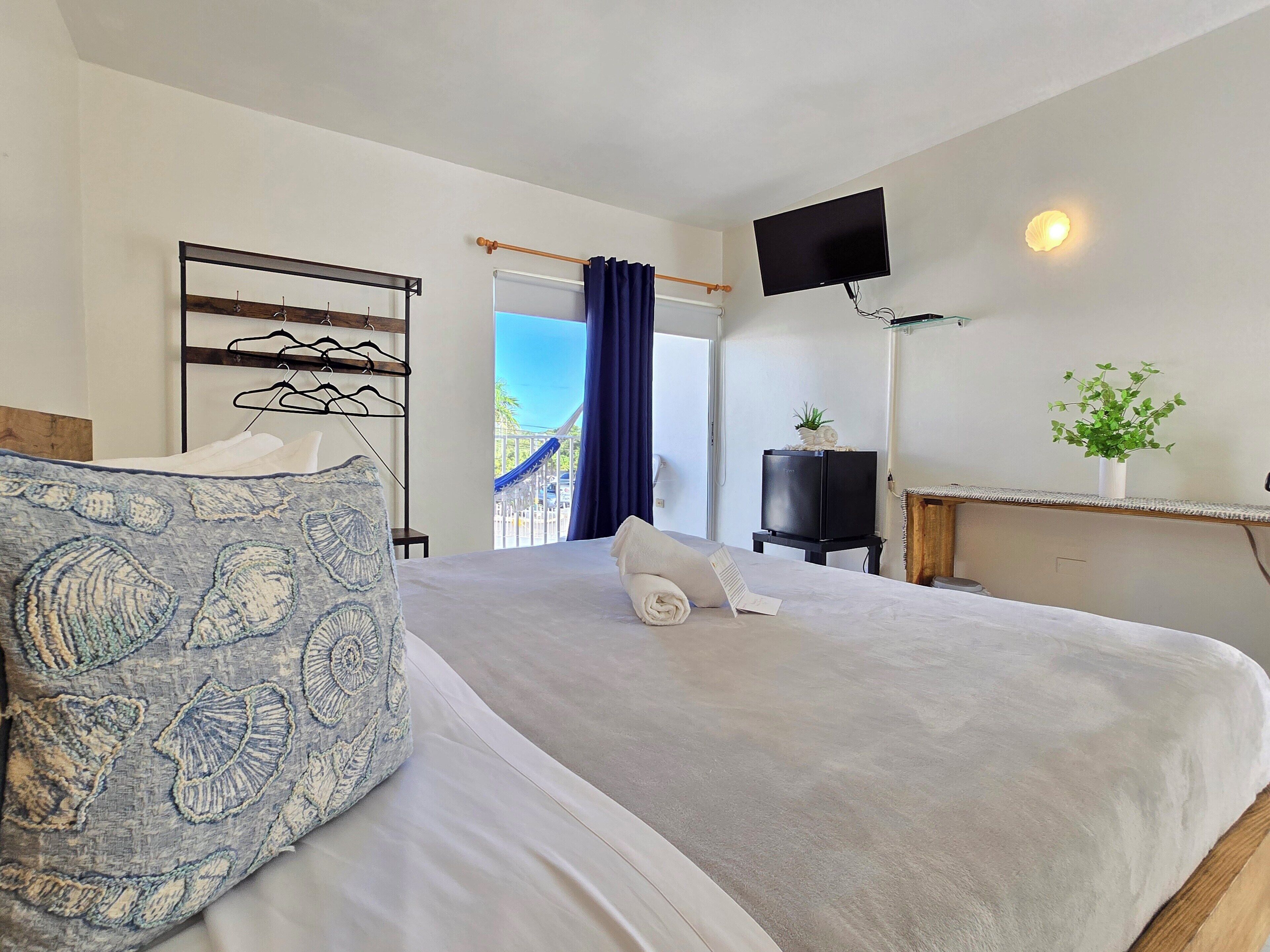 Ett hotellrum med en stor säng, en platt-TV som är monterad på väggen, en liten växt på ett sidobord och utsikt över omgivningarna genom en glasdörr.
