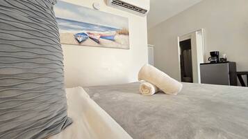 A hotel room with a bed, a painting of boats on a beach, an air conditioner, and a coffee machine.