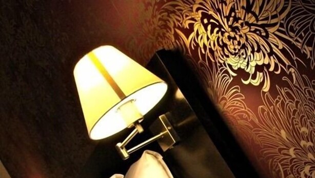 A lamp with a conical shade, a white pillow, and a wall with a floral pattern. - Hotel Belmonte (Norwich)