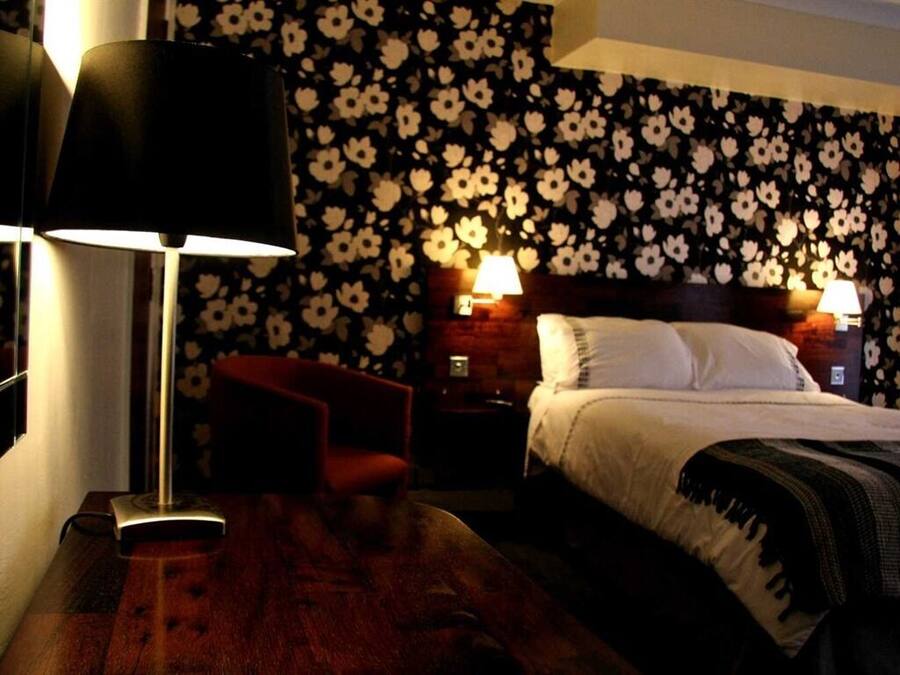 Quarto de hotel com uma cama, duas luminárias laterais, uma cadeira vermelha e piso de madeira.
