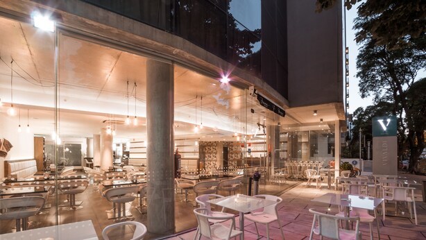 An outdoor cafe area with tables and chairs, a visible entrance to a building, and a sign with a 'V' logo. - Vivaldi Hotel Loft Punta Carretas (Montevideo)