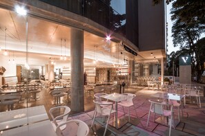 Un espace café en plein air avec des tables et des chaises, une entrée bien visible menant à un bâtiment, et une enseigne avec un logo en forme de « V ».