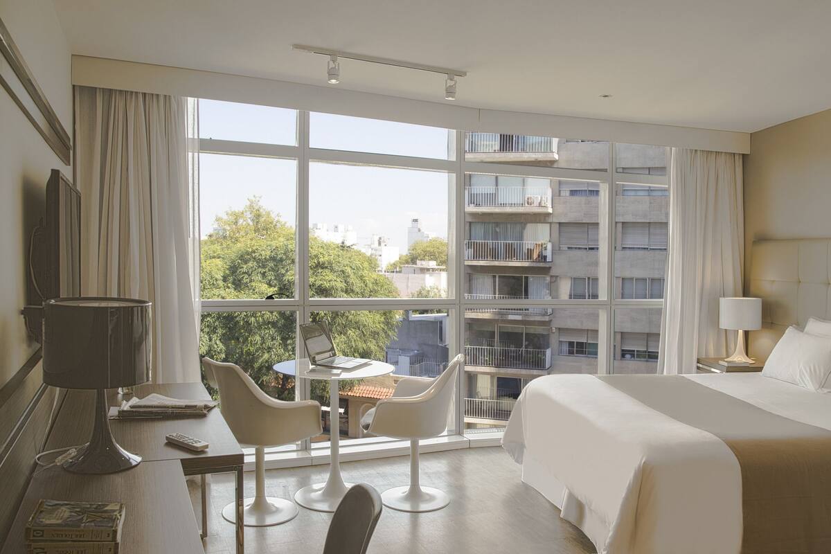 A hotel room with a large bed, a desk with a laptop, two chairs, and a view of a cityscape through large windows.