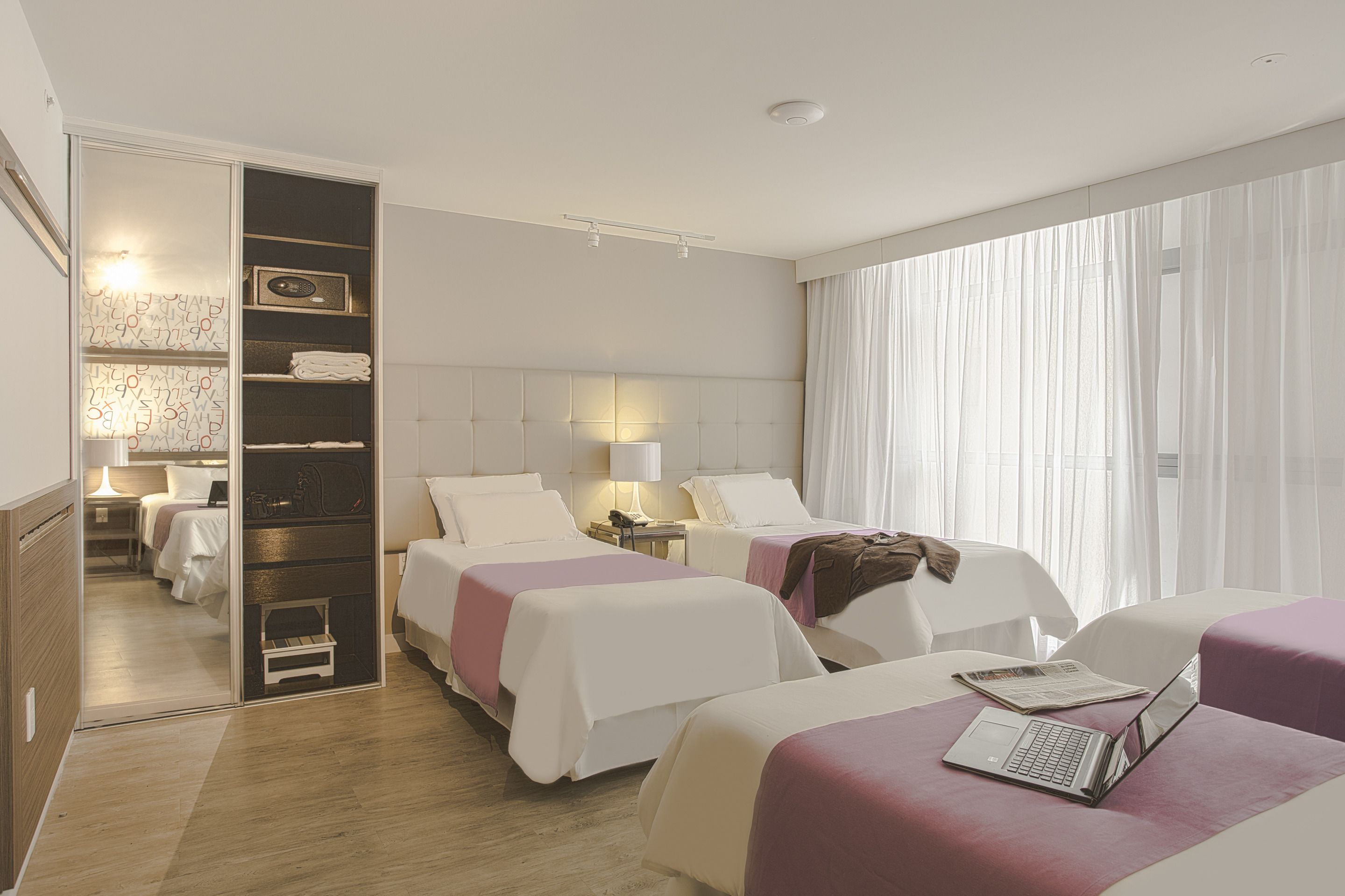 Ett hotellrum med två sängar, en rymlig klädkammare, ett skrivbord och ett fönster med tunna, genomskinliga gardiner.