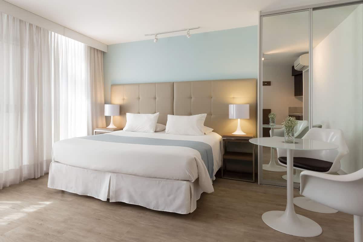 A modern hotel room with a large bed, two bedside tables with lamps, a glass table with a chair, and a large window with sheer curtains.