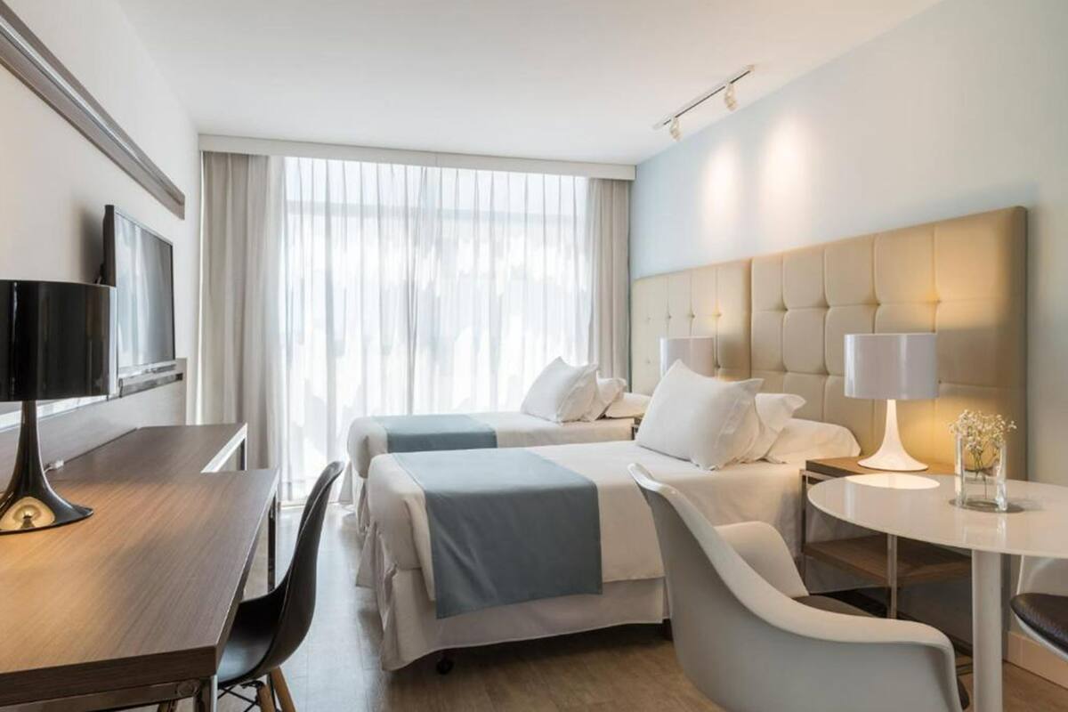 A hotel room with two beds, a desk with a TV, a round table with a lamp, and a large window with curtains.