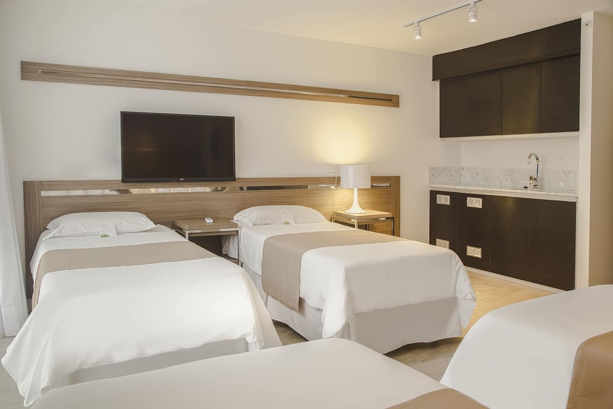 A hotel room with three single beds, a TV mounted on the wall, a bedside table with a lamp, and a kitchenette area with cabinets and a countertop.