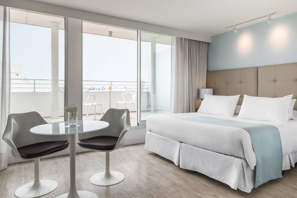 A modern hotel room with a large bed, a glass table with two chairs, and a balcony with a view.