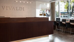 A reception desk with a wooden finish and a nameplate reading "VIVALDI". There are black chairs and a table with a computer monitor in the background. - Vivaldi Hotel Loft Punta Carretas (Montevideo)