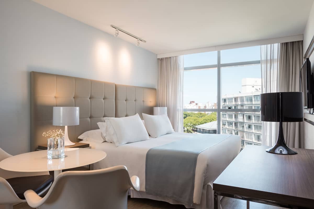 A hotel room with a large bed, a round table, and a view of the city.