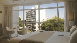 A hotel room with a large bed, a desk with a chair, a small table with a cup and a plate, and a view of buildings and trees through large windows. - Vivaldi Hotel Loft Punta Carretas (Montevideo)