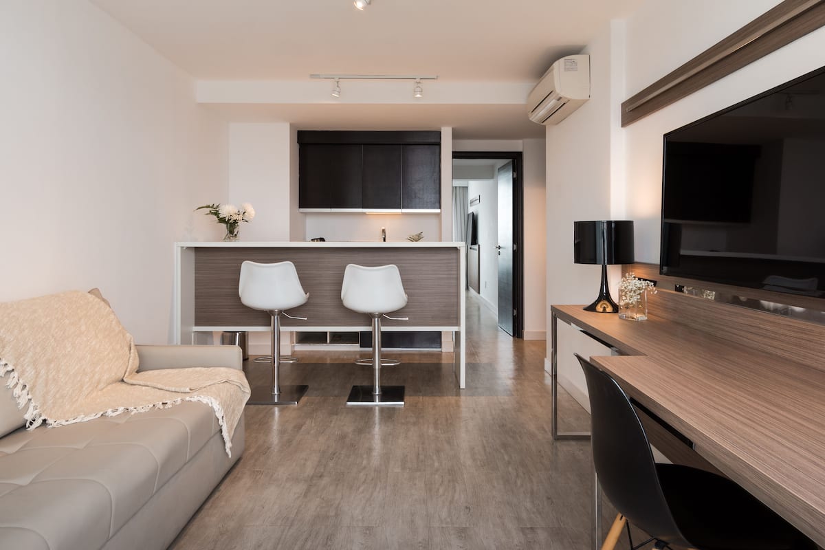 A modern living room with a sofa, a wooden dining table, two white bar stools, and a kitchen area in the background.