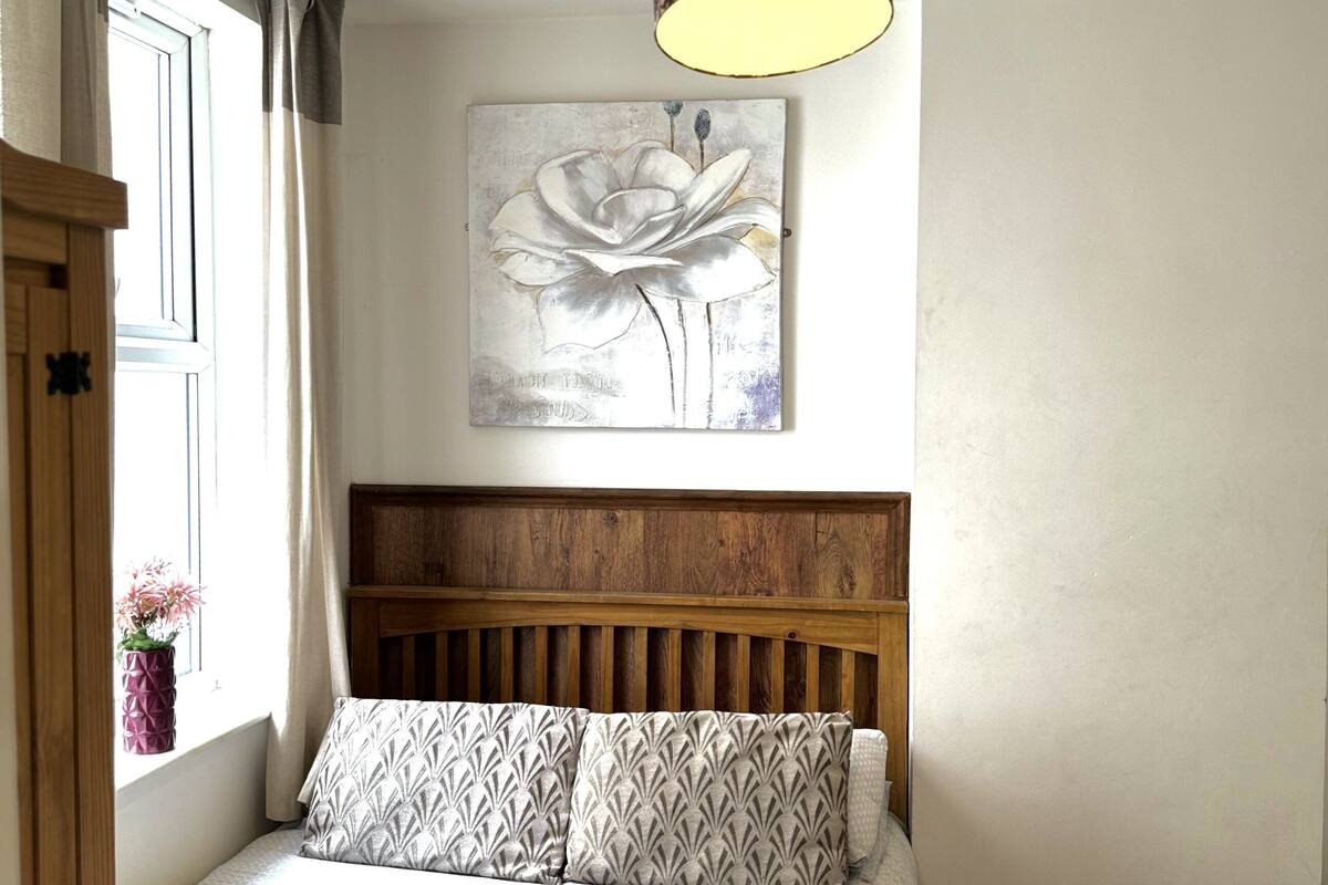 Une chambre à coucher avec une tête de lit en bois, un couvre-lit blanc, des coussins à motifs, une lampe fixée au mur et une fenêtre avec des rideaux.