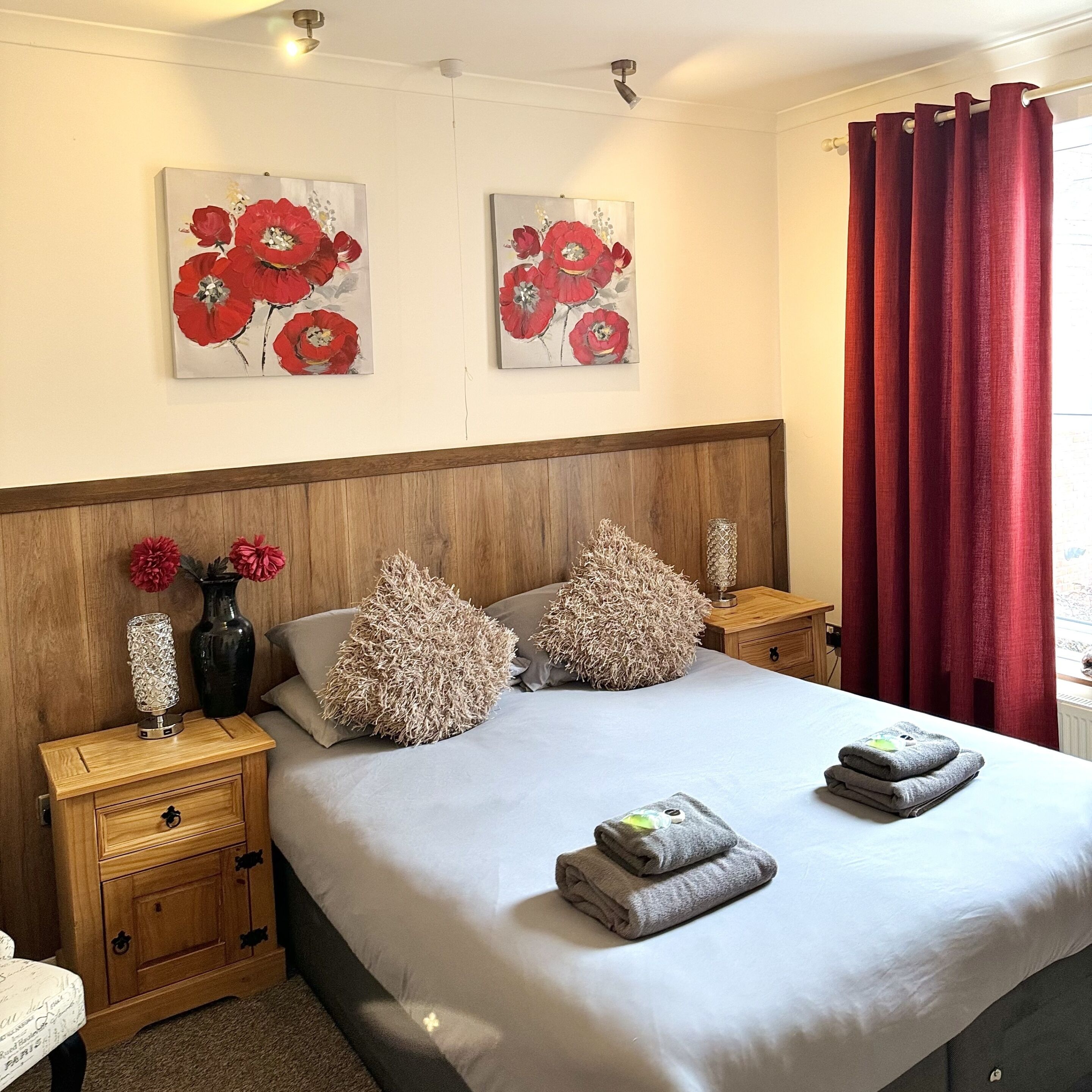 A neatly made bed with pillows and folded towels, wooden nightstands with drawers, and two floral paintings on the wall.