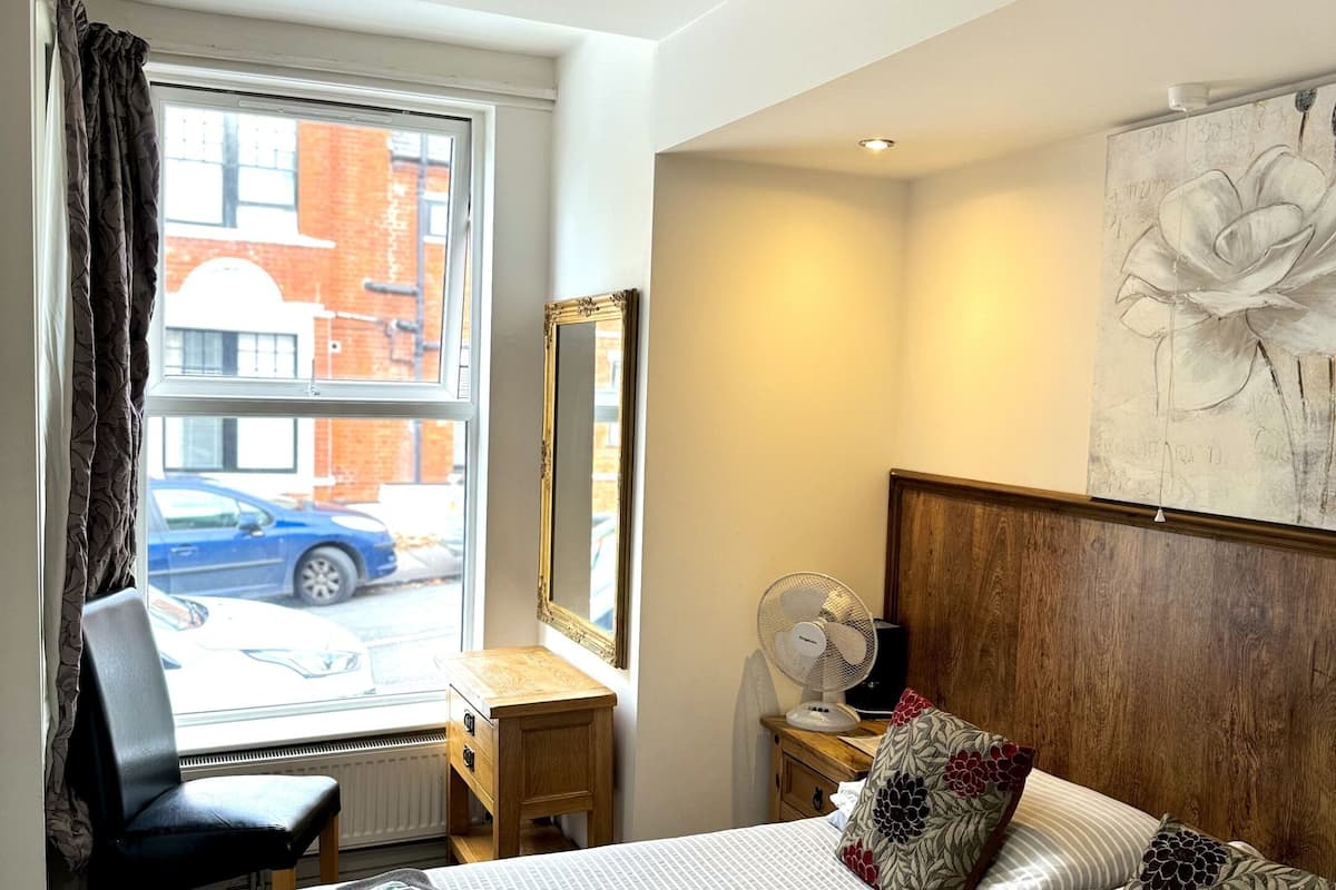 Une chambre d’hôtel avec un lit, une chaise, un bureau, un miroir, un ventilateur et une fenêtre donnant sur la rue.