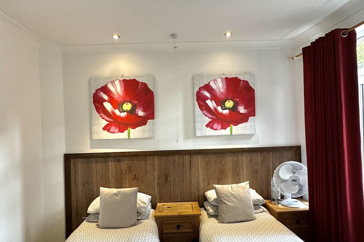Une chambre d’hôtel avec deux lits, une tête de lit en bois, une table de chevet avec un ventilateur et des œuvres d’art représentant des fleurs rouges accrochées au mur.
