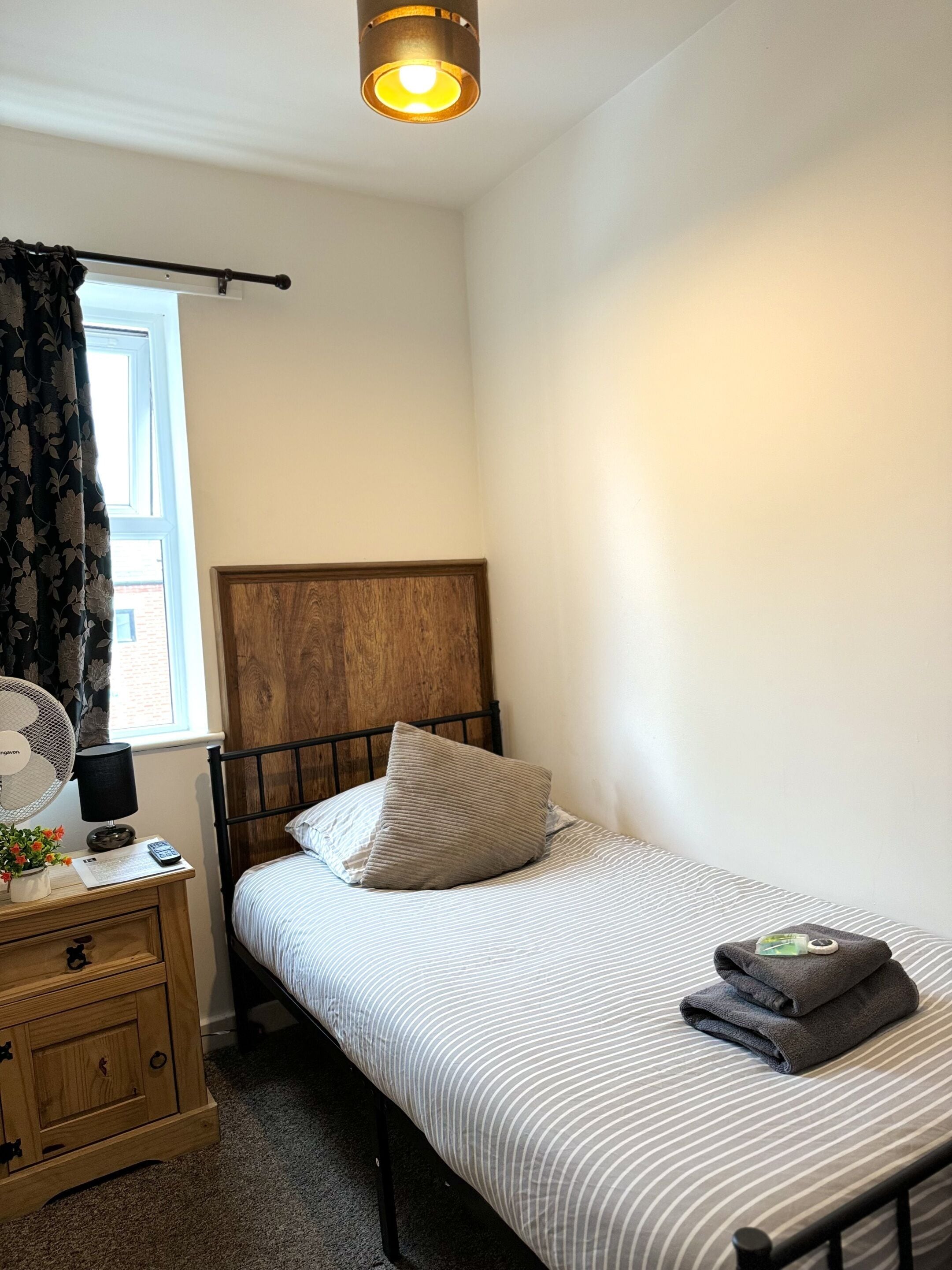 A bedroom with a single bed, a wooden headboard, a bedside table with a lamp, a fan, and a window with curtains.
