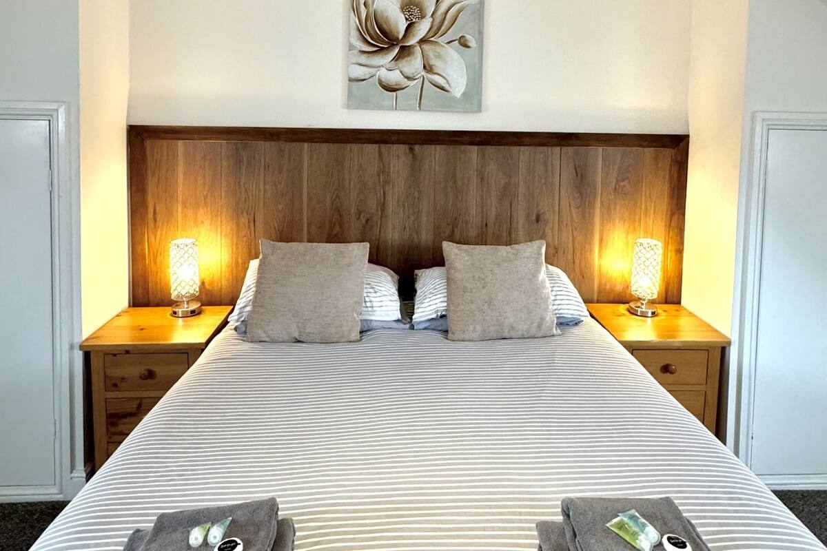 Une chambre d’hôtel avec un grand lit, deux tables de chevet avec des lampes, une tête de lit en bois et une œuvre d’art florale au-dessus du lit.