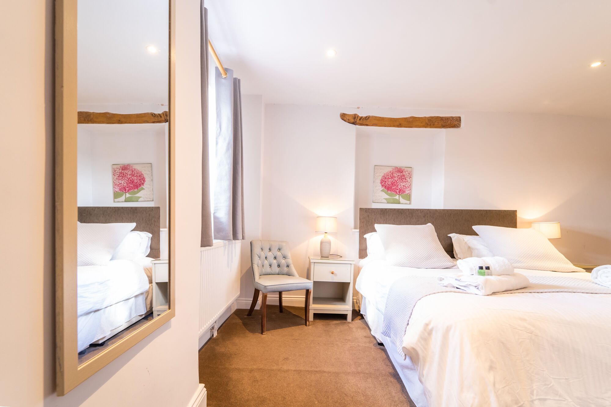 A hotel room with two beds, a chair, a nightstand, a mirror, and artwork on the walls.