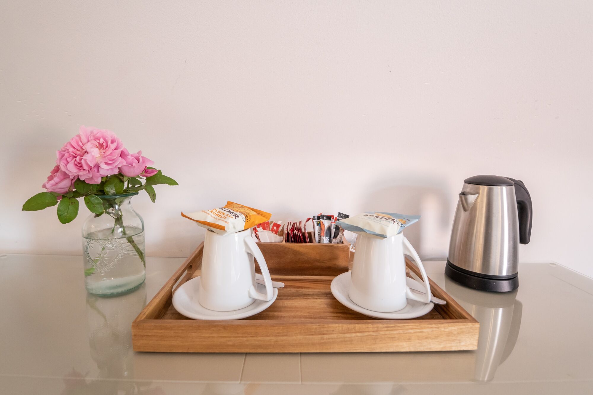 Una safata amb dues tasses de cafè blanques, una cafetera d'acer inoxidable i un ram de flors roses sobre una superfície reflectant.