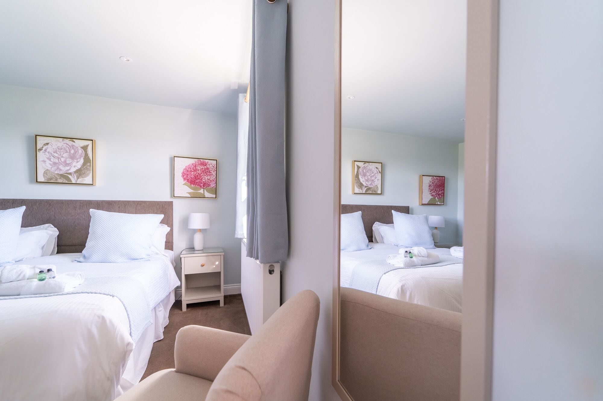 A hotel room with two beds, a bedside table with a lamp, and floral artwork on the walls.