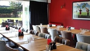 Un restaurant avec de longues tables en bois, des chaises à revêtement gris et un mur d’accent rouge orné d’un tableau.