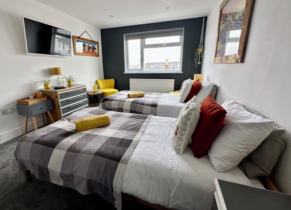 A modern bedroom with two beds, a desk, a chair, and a window with a view.