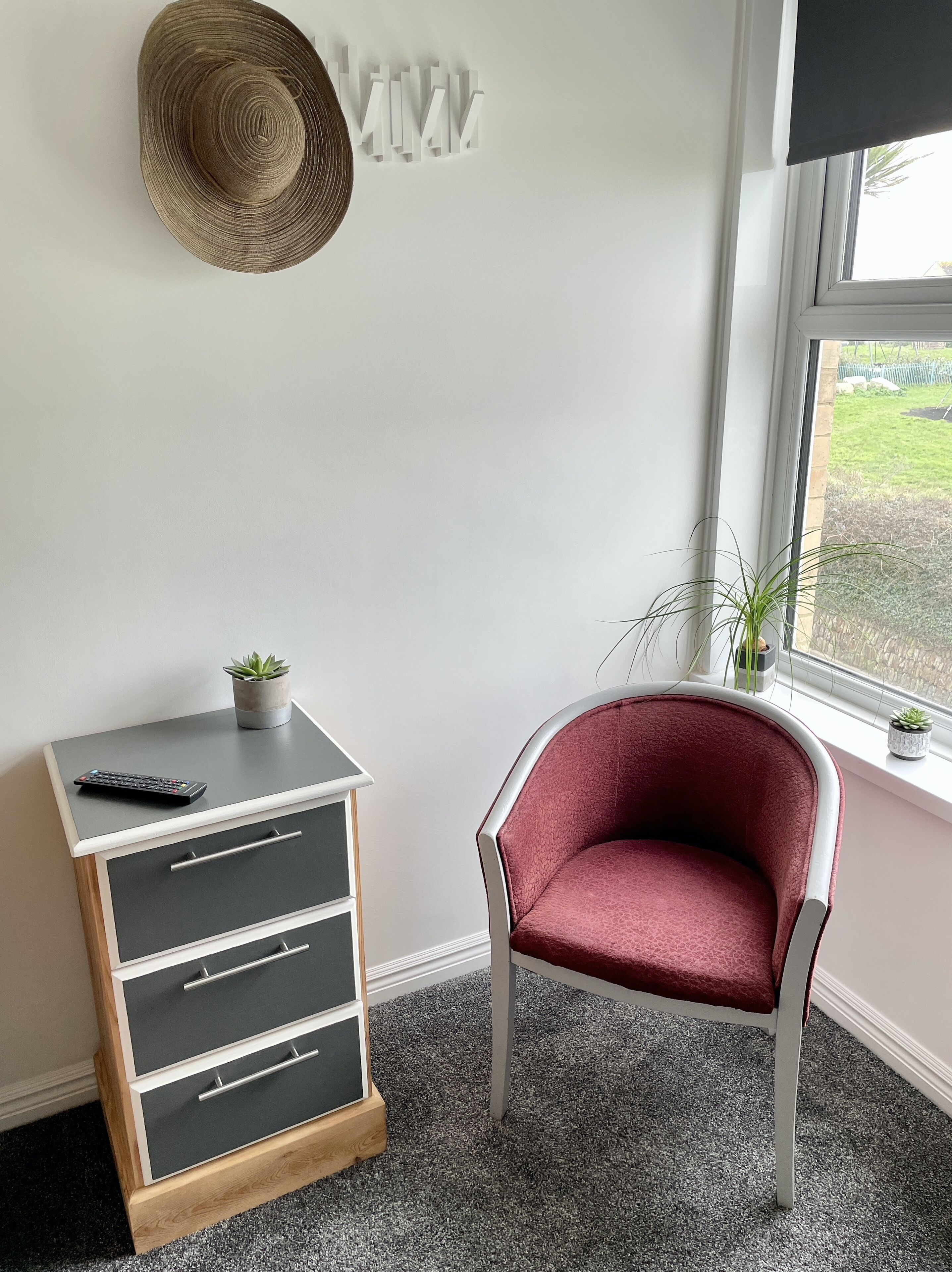 Ein Zimmer mit einem Holzkasten, einem Schubladenschrank, einem roten Stuhl, einem Strohhut an der Wand und einem Fenster mit Blick auf Grünflächen.