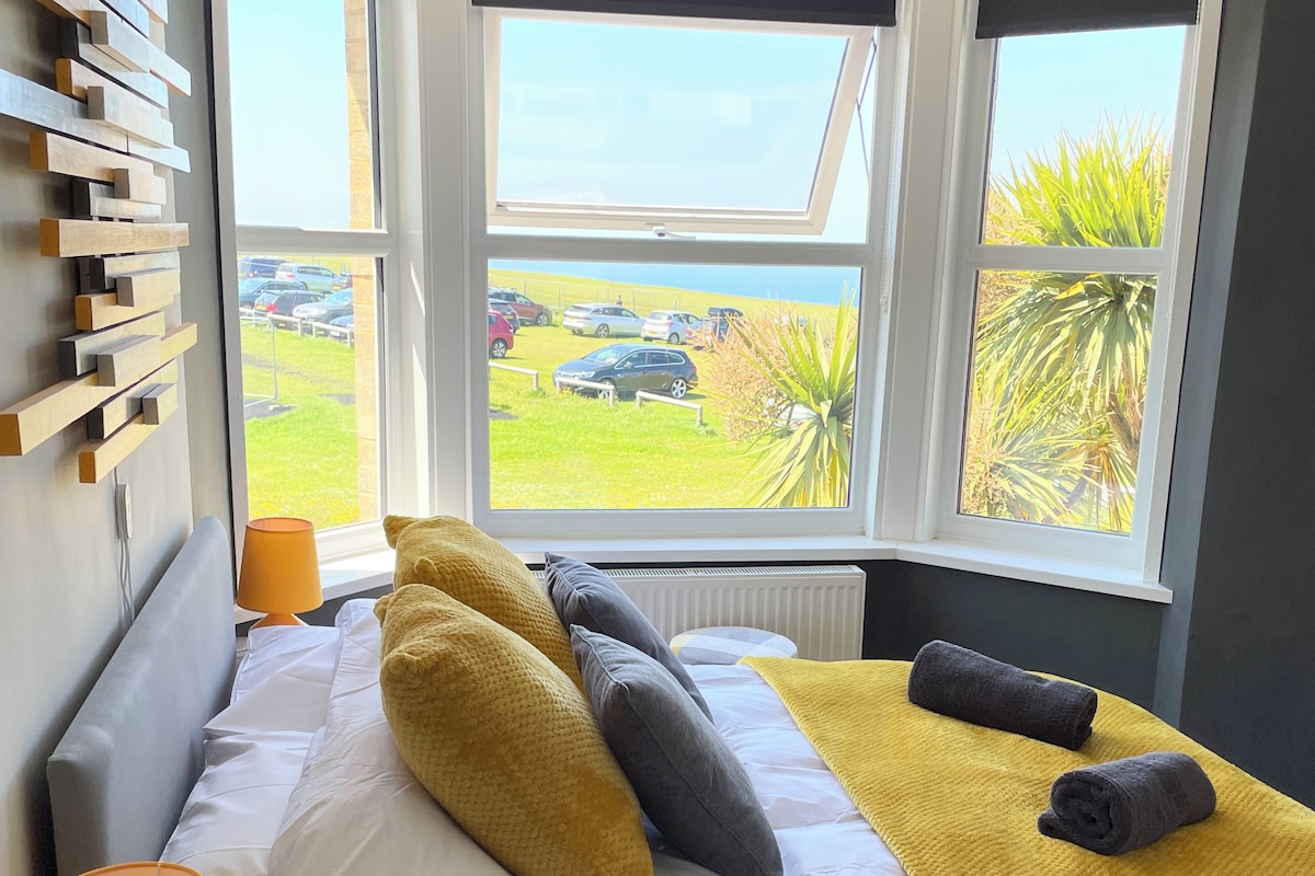 Un dormitorio con cama, almohadas, una lámpara de noche y vistas a un estacionamiento a través de amplias ventanas.
