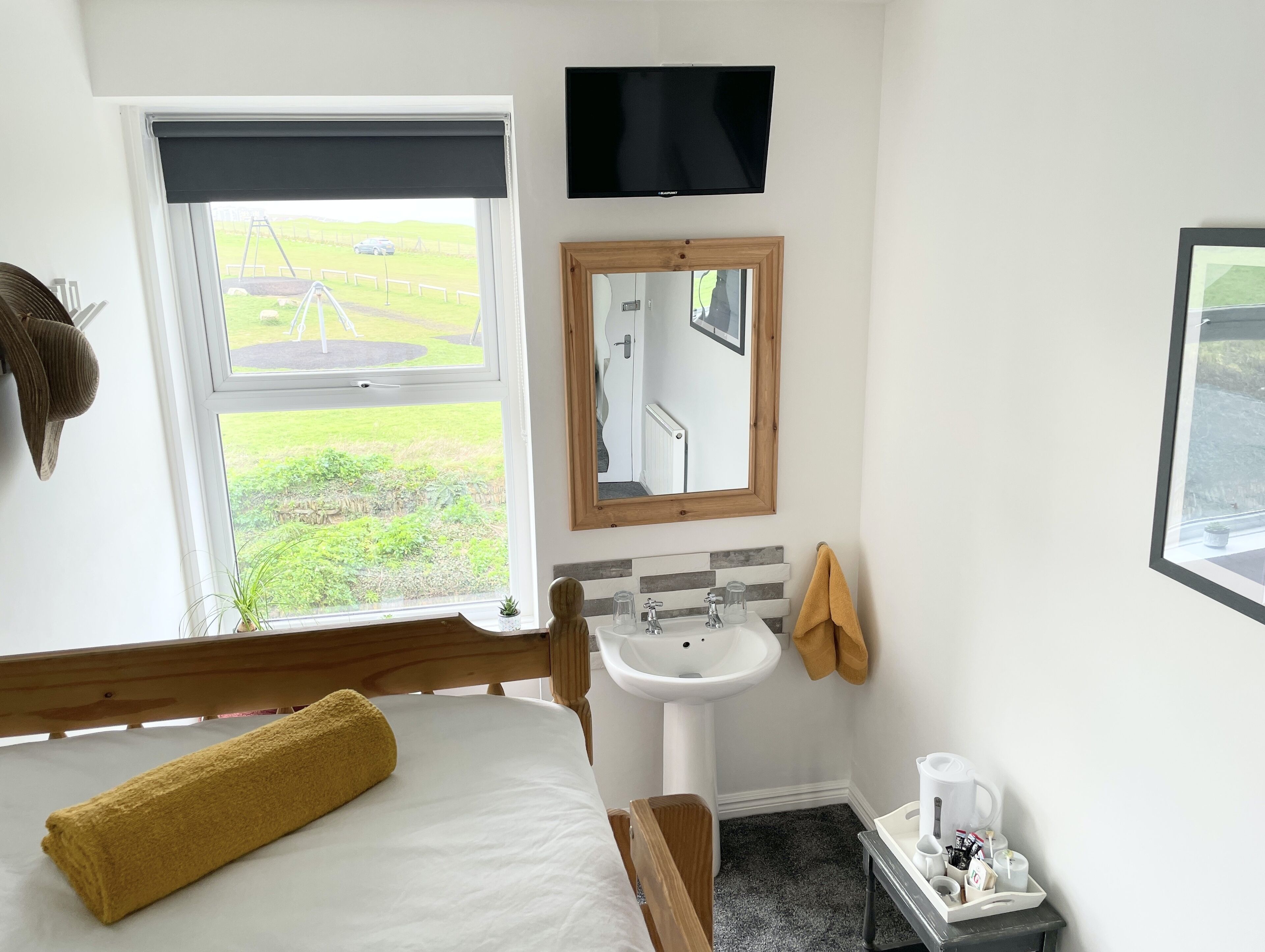 A hotel room with a bed, a window with a view of a grassy area, a sink, a mirror, and a television.