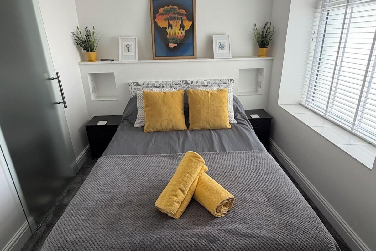 Un dormitorio con una cama, dos mesitas de noche, una ventana con persianas, un cuadro en la pared y una planta en maceta sobre las mesitas de noche.