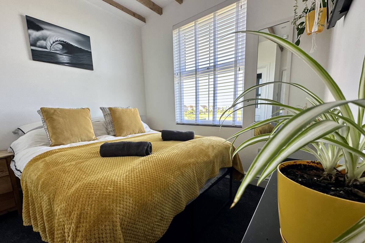 Un lit bien fait, recouvert d’une couverture jaune, avec deux oreillers et une serviette enroulée. Une grande fenêtre avec des stores, une plante en pot et un tableau ornent le mur.