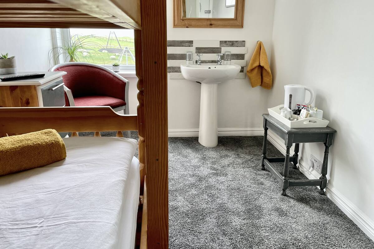 Une chambre avec un lit superposé, une table de chevet avec une lampe, une chaise rouge, une fenêtre donnant sur une balançoire et une salle de bain avec un lavabo, un miroir et un porte-serviettes.