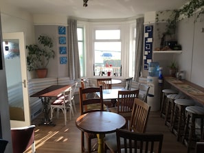 A dining area with a round table and chairs, a kitchen counter with stools, and a view through large windows. - Breakers Guest House (Newquay)
