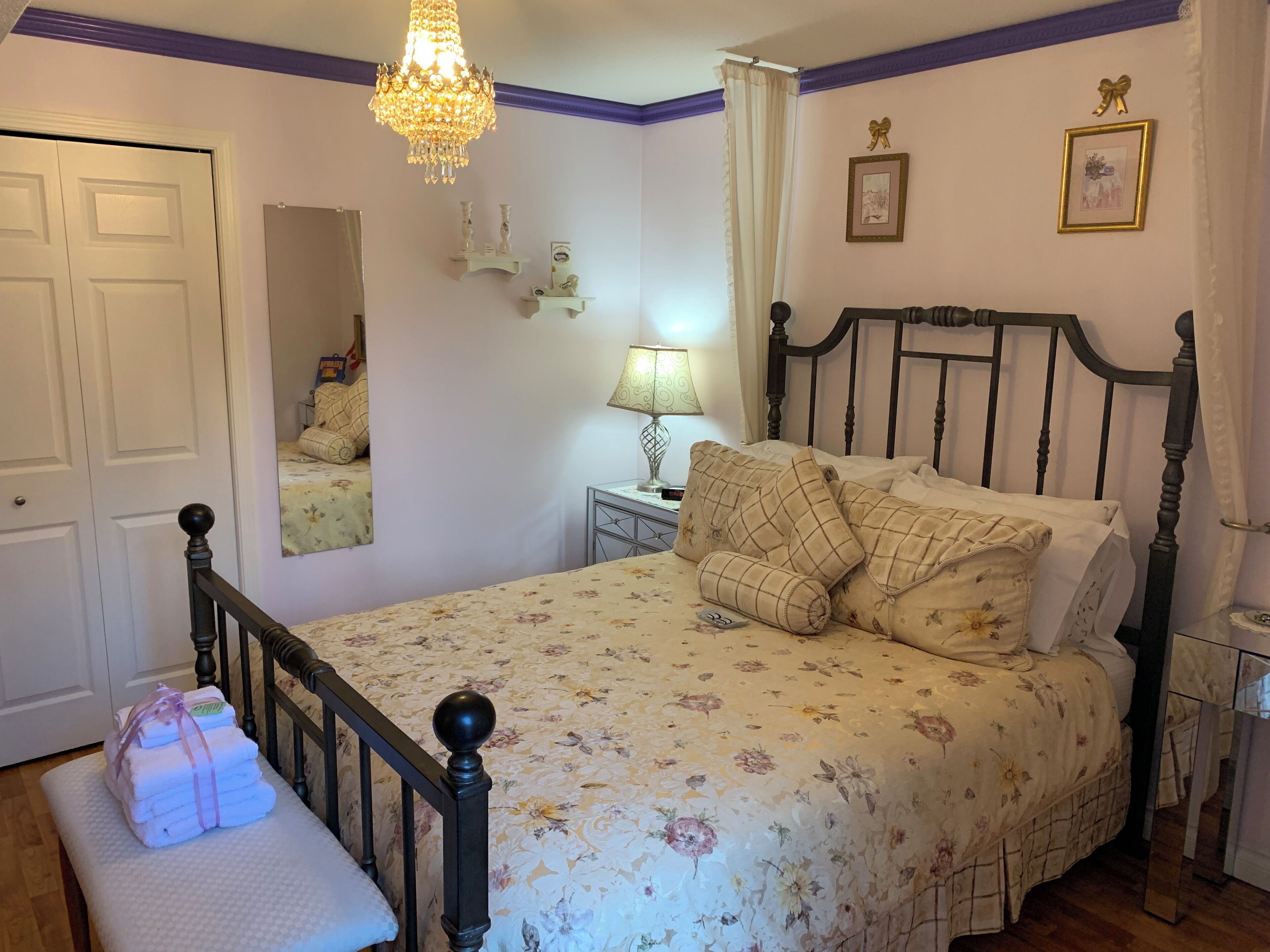 Quarto com colcha florida, cabeceira de madeira, luminária de cabeceira, espelho e pilha de toalhas em um banco.