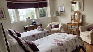 Une chambre d’hôtel avec deux lits, un bureau avec une lampe, une chaise, une coiffeuse avec un miroir et une fenêtre donnant sur un paysage verdoyant.