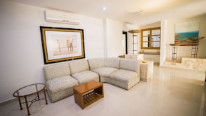Uma sala de estar com sofá seccional, uma mesinha de centro, um criado-mudo e um quadro na parede.