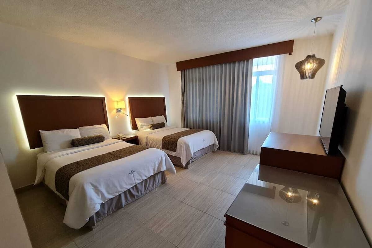 A hotel room with two beds, a desk, a TV, and a window with curtains.