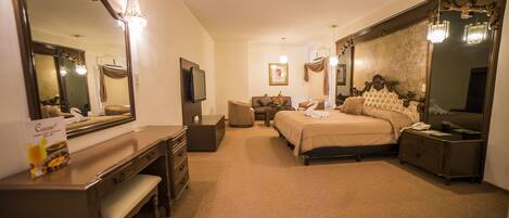 A hotel room with a large bed, a desk with a mirror, a dresser, a bedside table, a TV, and a chandelier.