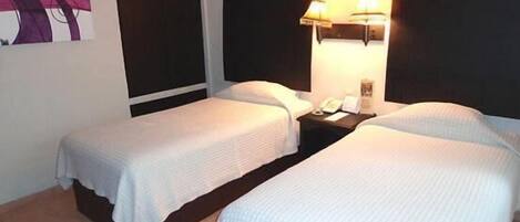 Camera d'albergo con due letti singoli, un comodino con lampada e una TV a parete.