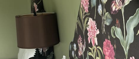 Una cama con sábanas blancas, un cabecero floral, una lámpara oscura y un control remoto sobre una mesita de noche de madera.