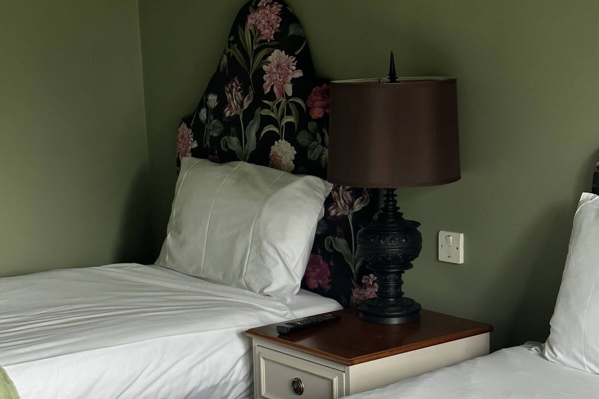Habitación de hotel con dos camas, una mesita de noche con lámpara y cabecera estampada con motivos florales.