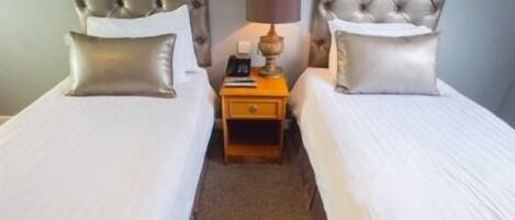 A twin room with two beds, each with a tufted headboard, a bedside table with a lamp, and a small drawer.