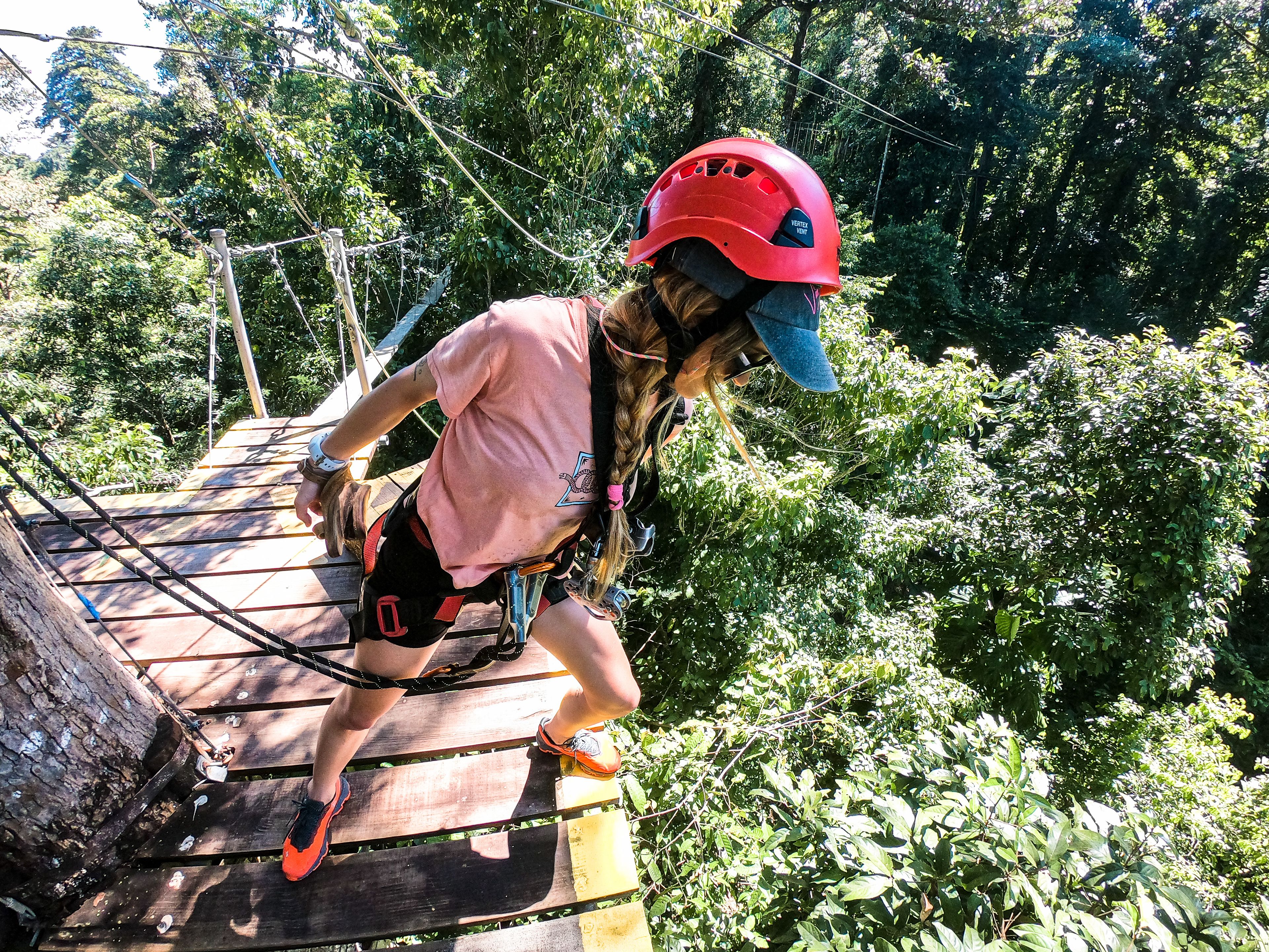 A person on a wooden bridge with safety harnesses, surrounded by dense greenery.
