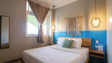 A hotel room with a bed, a window with curtains, a wall-mounted macrame piece, and a bedside table with a lamp.