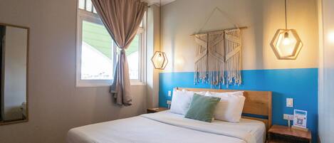 Een hotelkamer met een bed, een raam met gordijnen, een macramé wanddecoratie en een nachtkastje met een lamp.