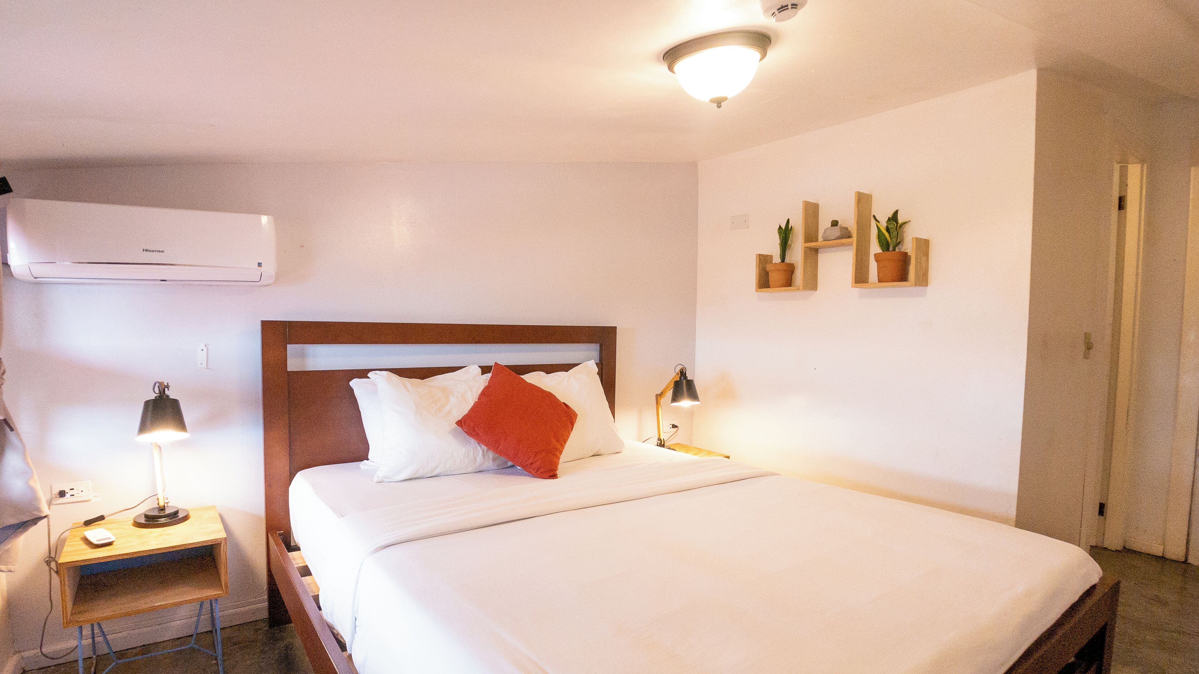 A hotel room with a bed, bedside lamps, a small desk, and a wall-mounted air conditioner.