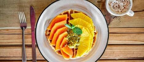 A plate of waffles topped with sliced mango and pineapple, a mint leaf, and passion fruit sauce, accompanied by a cup of coffee on a wooden table.