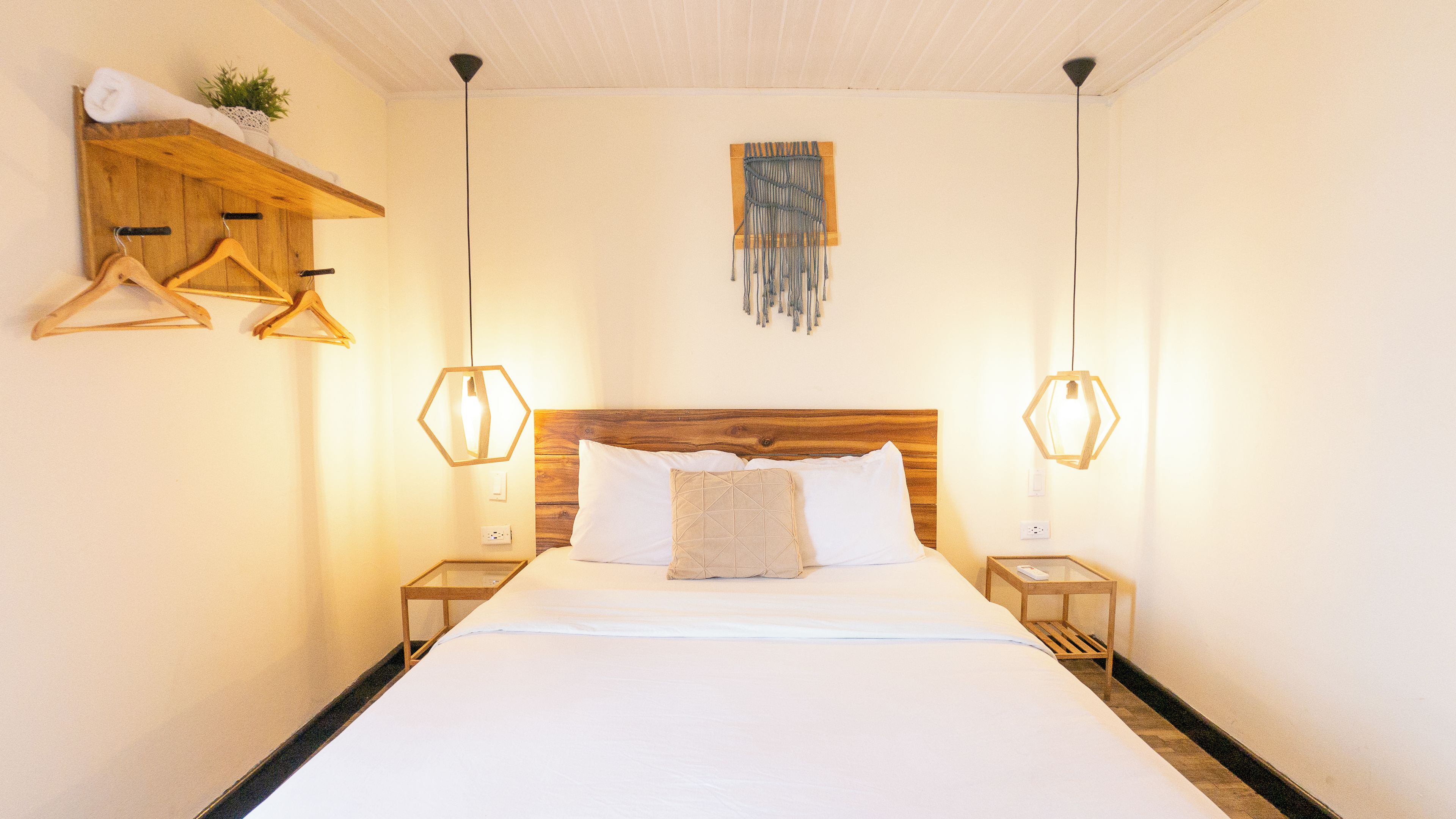 Una habitación de hotel con cabecera de madera, cama con ropa blanca, dos mesitas de noche con lámparas y un estante de madera con una toalla y una planta.